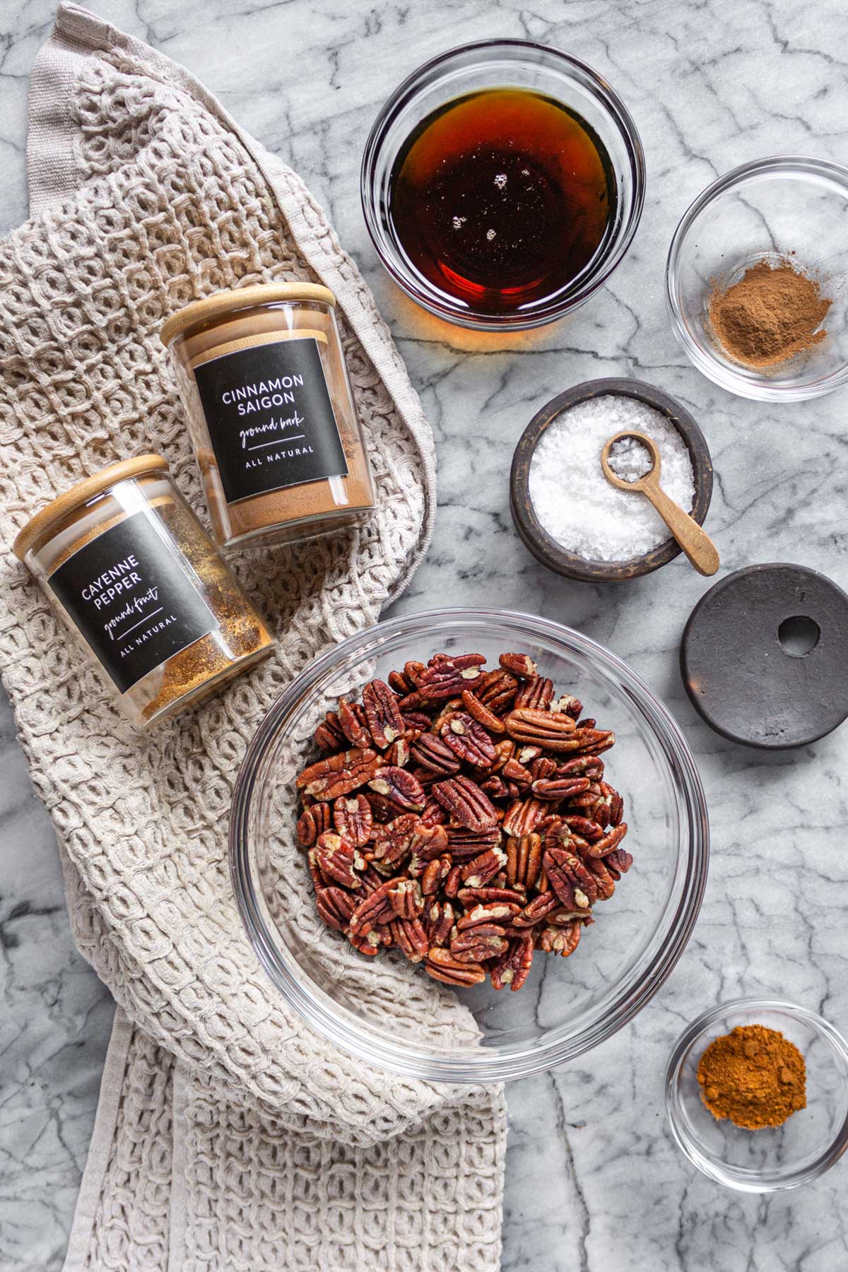 The ingredients for homemade Maple Pecans in glass bowls on a marble board with a beige waffle knit kitchen towel: Pecans, Maple Syrup, Cinnamon, Cayenne Pepper.