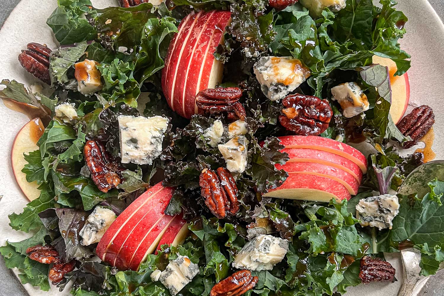 A close up of a plate filled with a kale salad with apples, blue cheese crumbles, and candied maple pecans.