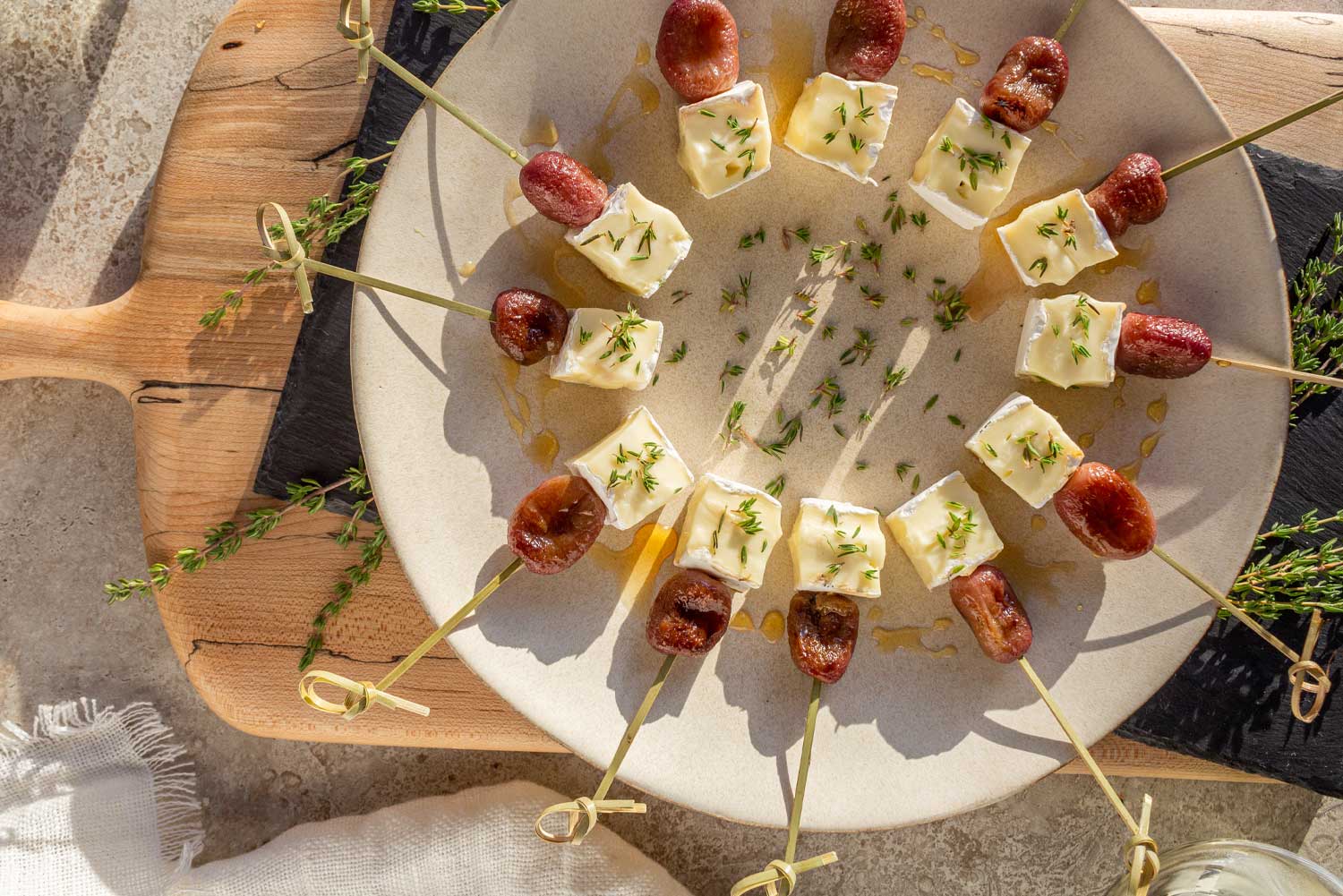 Another view of a round platter in the late afternoon sunlight holding Roasted Grape & Brie appetizer bites on skewers. Cubes of brie and roasted grapes are on natural skewers and drizzled with honey with fresh thyme scattered on the plate.