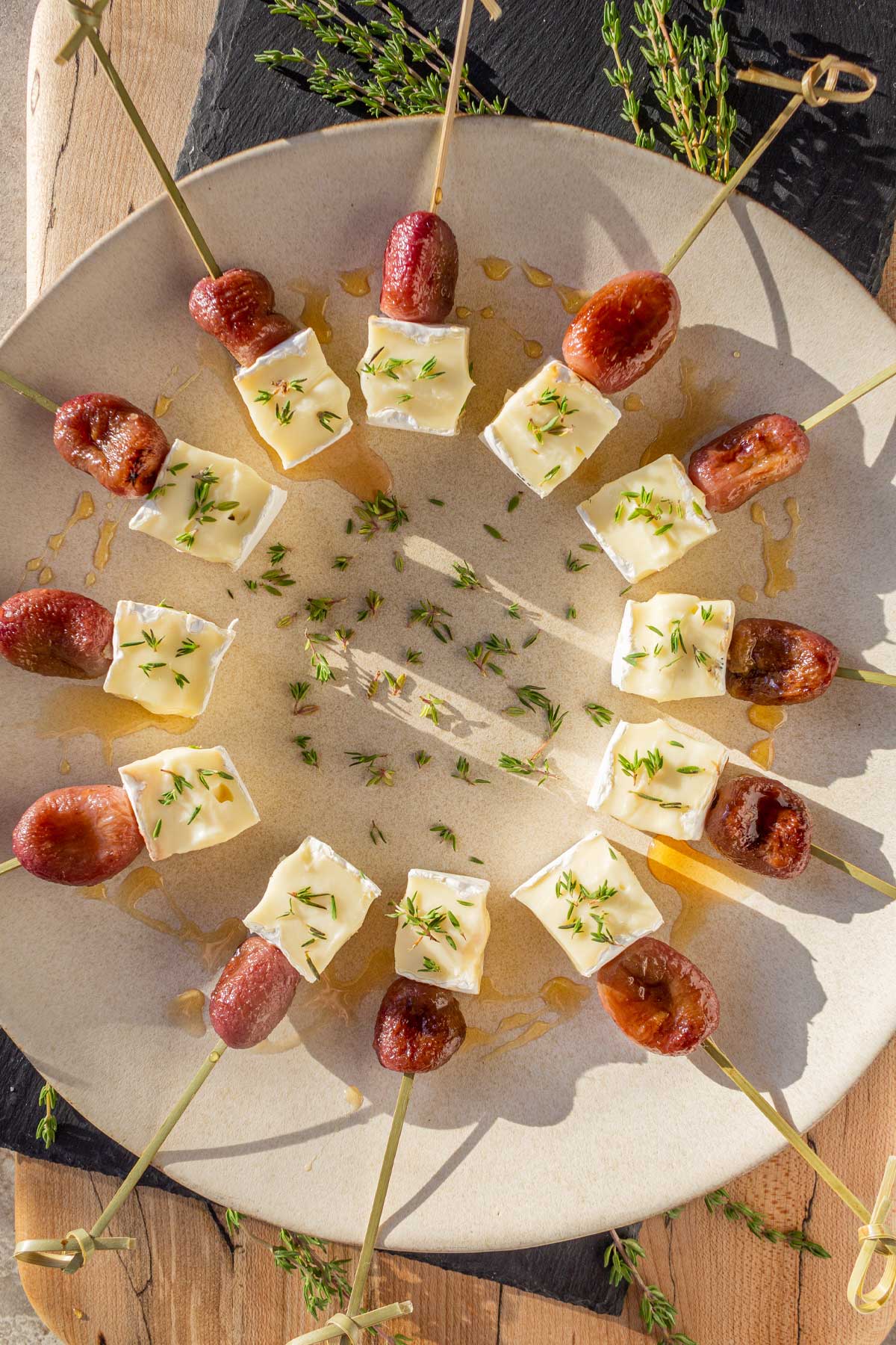 Another view of a round platter in the late afternoon sunlight holding Roasted Grape & Brie appetizer bites on skewers. Cubes of brie and roasted grapes are on natural skewers and drizzled with honey with fresh thyme scattered on the plate.