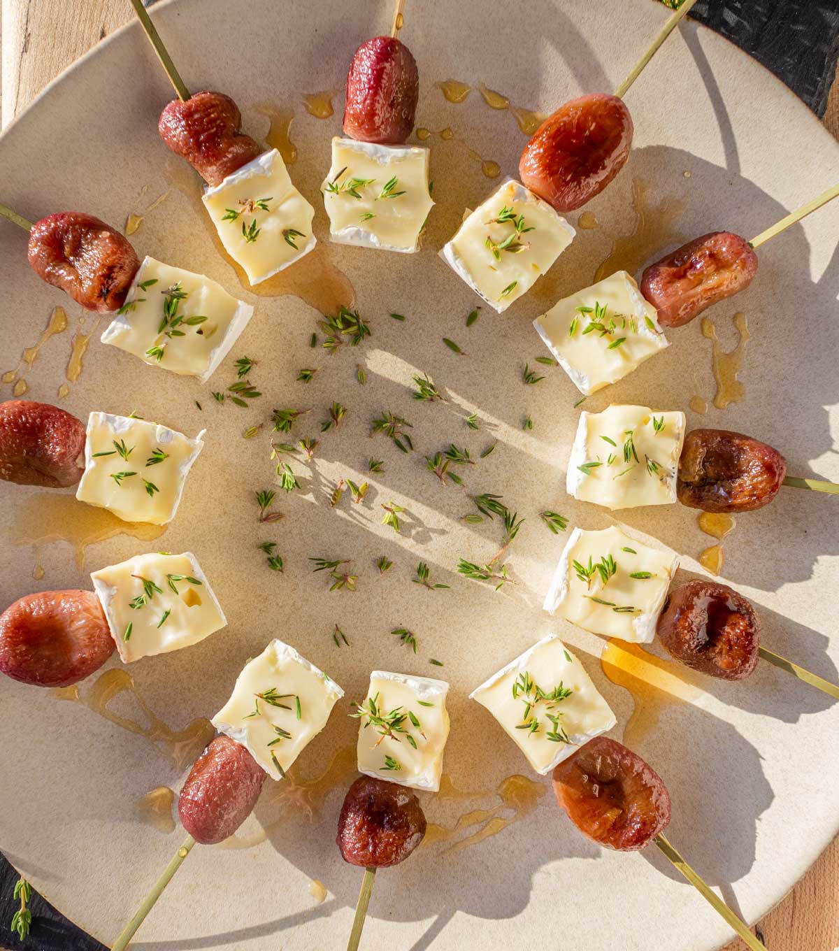 A platter filled with appetizer skewers of brie and roasted grapes drizzled in honey and sprinkled with fresh thyme.