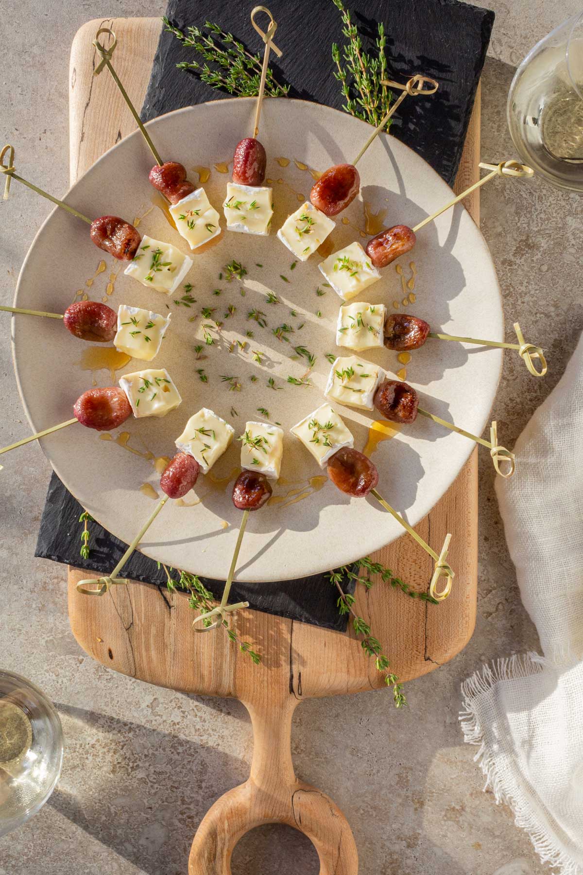 A round platter in the late afternoon sunlight holding Roasted Grape & Brie appetizer bites on skewers. Cubes of brie and roasted grapes are on natural skewers and drizzled with honey with fresh thyme scattered on the plate.