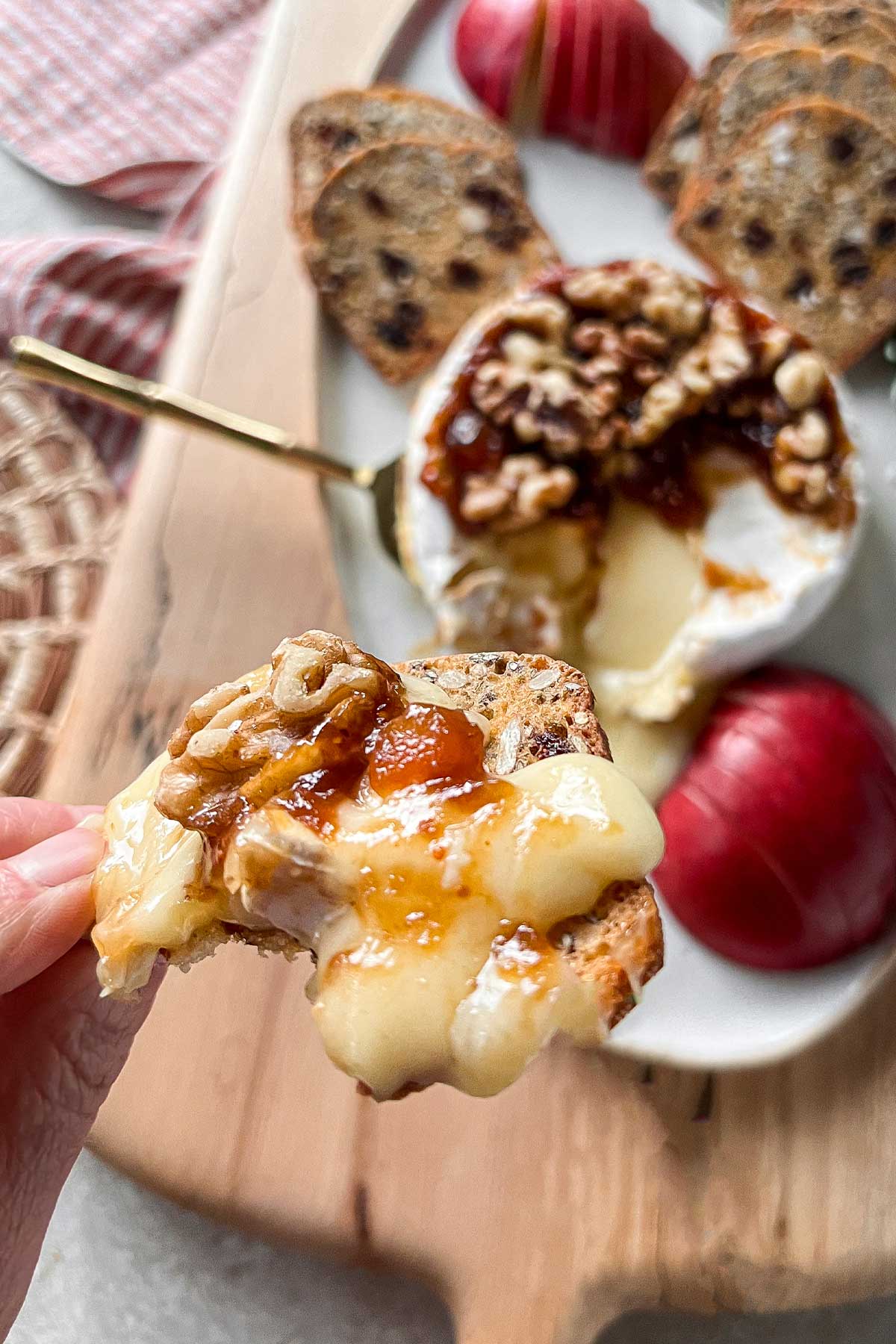 A close up of a cracker topped with melted Baked Brie And Preserves with a walnut on top. In the background, is a cheese board with a Baked Brie, sliced apples, and crackers.