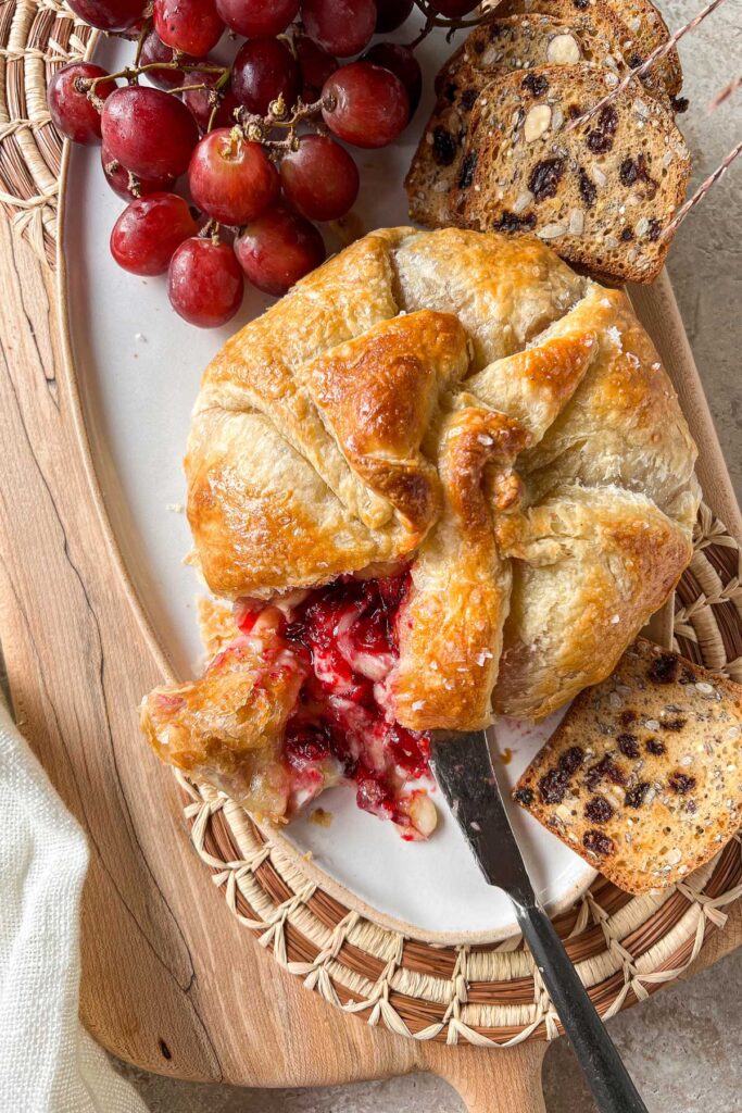 A ceramic platter on top of a maple serving board with a fresh from the oven Cranberry Baked Brie En Croûte.  The brie is cut with cheese and cranberry sauce spilling out onto the platter.