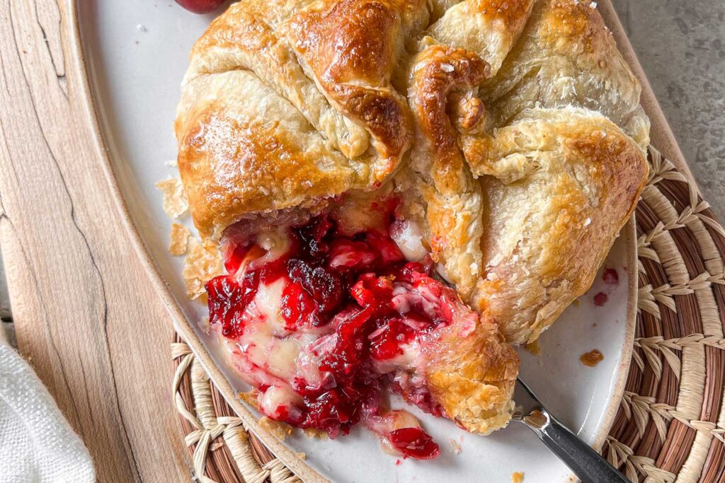 A ceramic platter on top of a maple serving board with a fresh from the oven Cranberry Baked Brie En Croûte.  The brie is cut with cheese and cranberry sauce spilling out onto the platter.