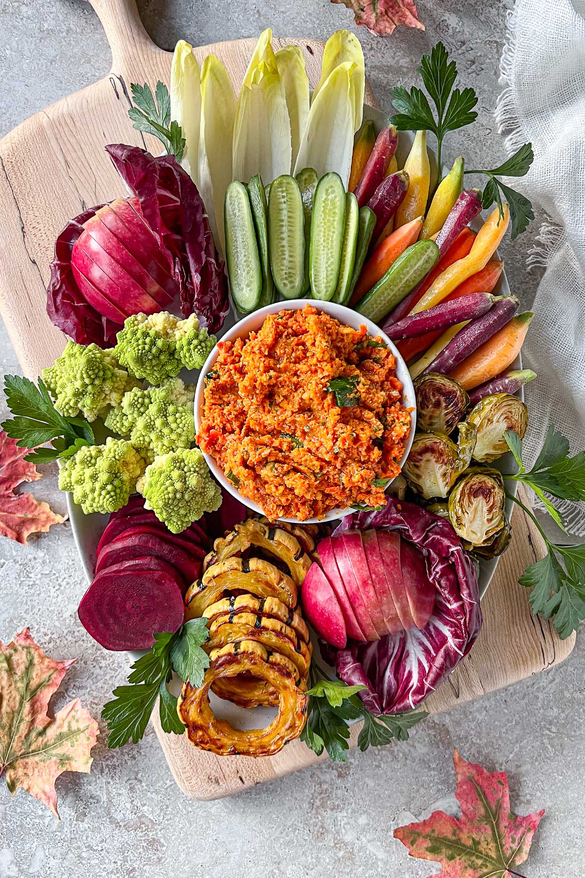 A bowl of bright orange Smoky Roasted Carrot Dip sitting on top of a maple serving board surrounded by a fresh vegetable crudité platter and almond, date crackers.