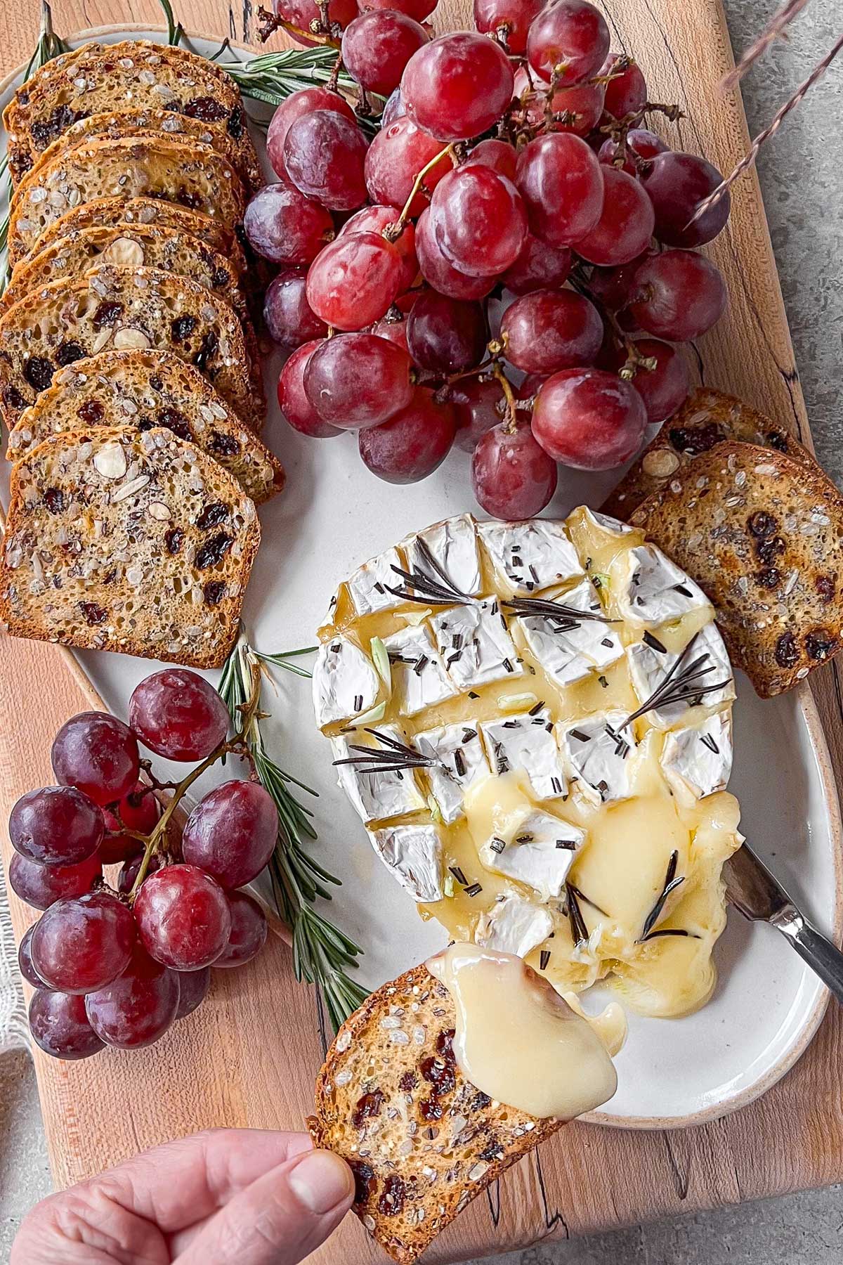 A spalted maple cheese board and ceramic platter filled with an oozing baked brie studded with garlic and rosemary. The cheese is surrounded by red grapes and crackers studded with dates and almonds.