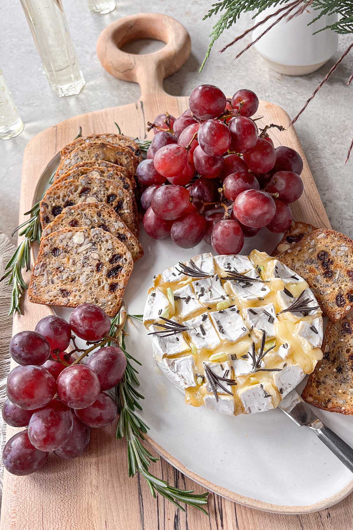 A spalted maple cheese board and ceramic platter filled with an oozing baked brie studded with garlic and rosemary. The cheese is surrounded by red grapes and crackers studded with dates and almonds.