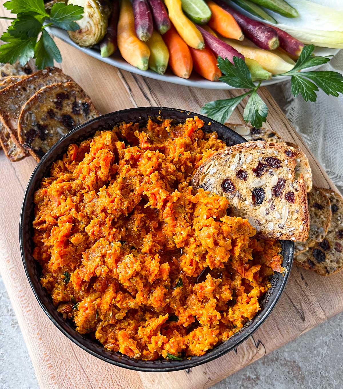 A pottery bowl on top of a maple serving board filled with a bright orange Smoky Roasted Carrot Dip.