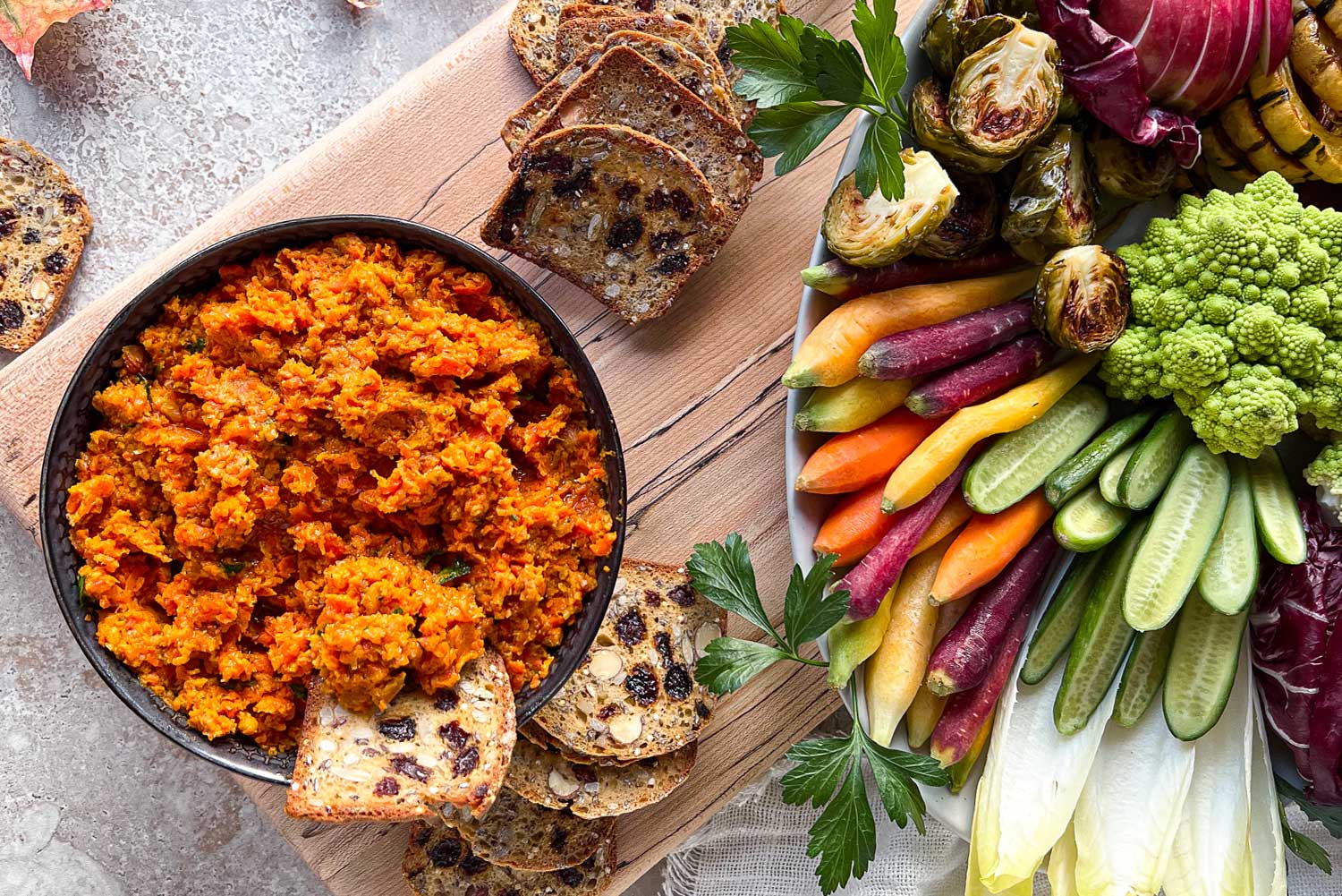 A bowl of bright orange Smoky Roasted Carrot Dip sitting on top of a maple serving board surrounded by a fresh vegetable crudité platter and almond, date crackers.
