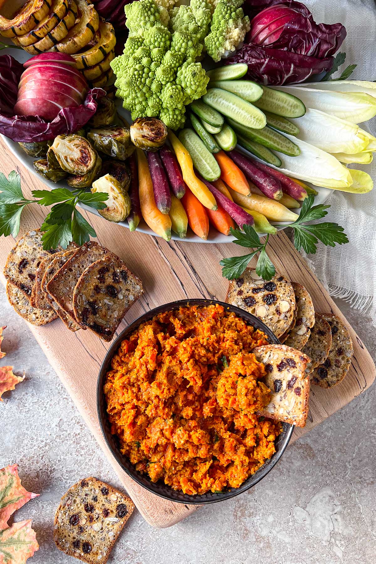 A bowl of bright orange Smoky Roasted Carrot Dip sitting on top of a maple serving board surrounded by a fresh vegetable crudité platter and almond, date crackers.
