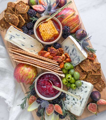 A Spalted Maple wooden cheese board filled with a tasting assortment of 3 blue cheeses. They are surrounded by fresh fruit, nuts, olives, crackers, jam, and honeycomb.