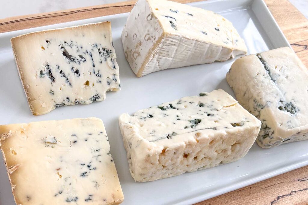 A white ceramic cheese platter showing the different types of blue cheese: Roquefort, Gorgonzola, Cambozola, Stilton, Domestic Blue