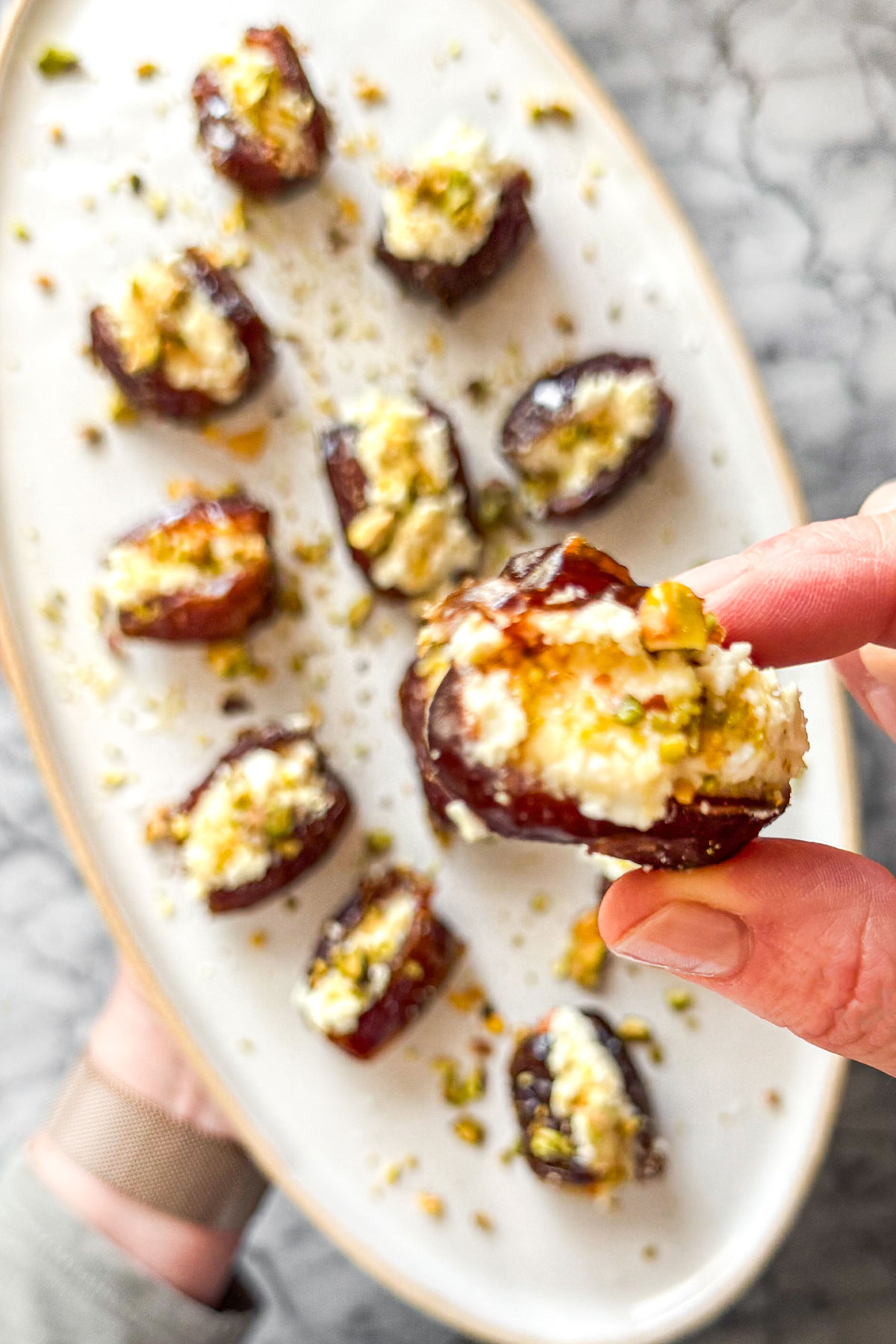 A white oval ceramic platter on a marble counter filled with Goat Cheese Stuffed Dates. The dates are drizzled with hot honey and topped with chopped pistachios which are scattered on the platter.