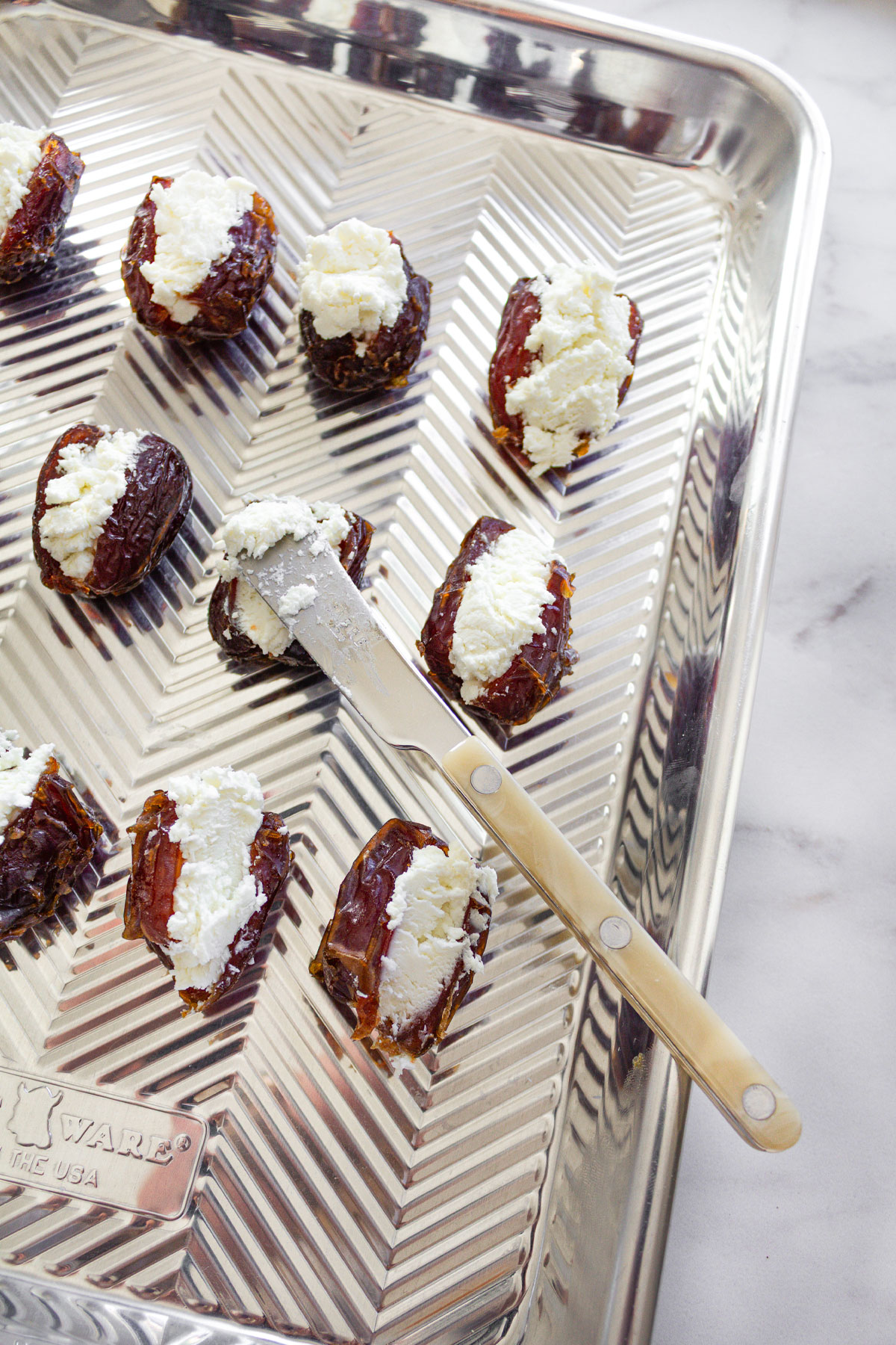 A baking sheet lined with Goat Cheese Stuffed Dates and a spreader spoon, waiting to go in the oven.