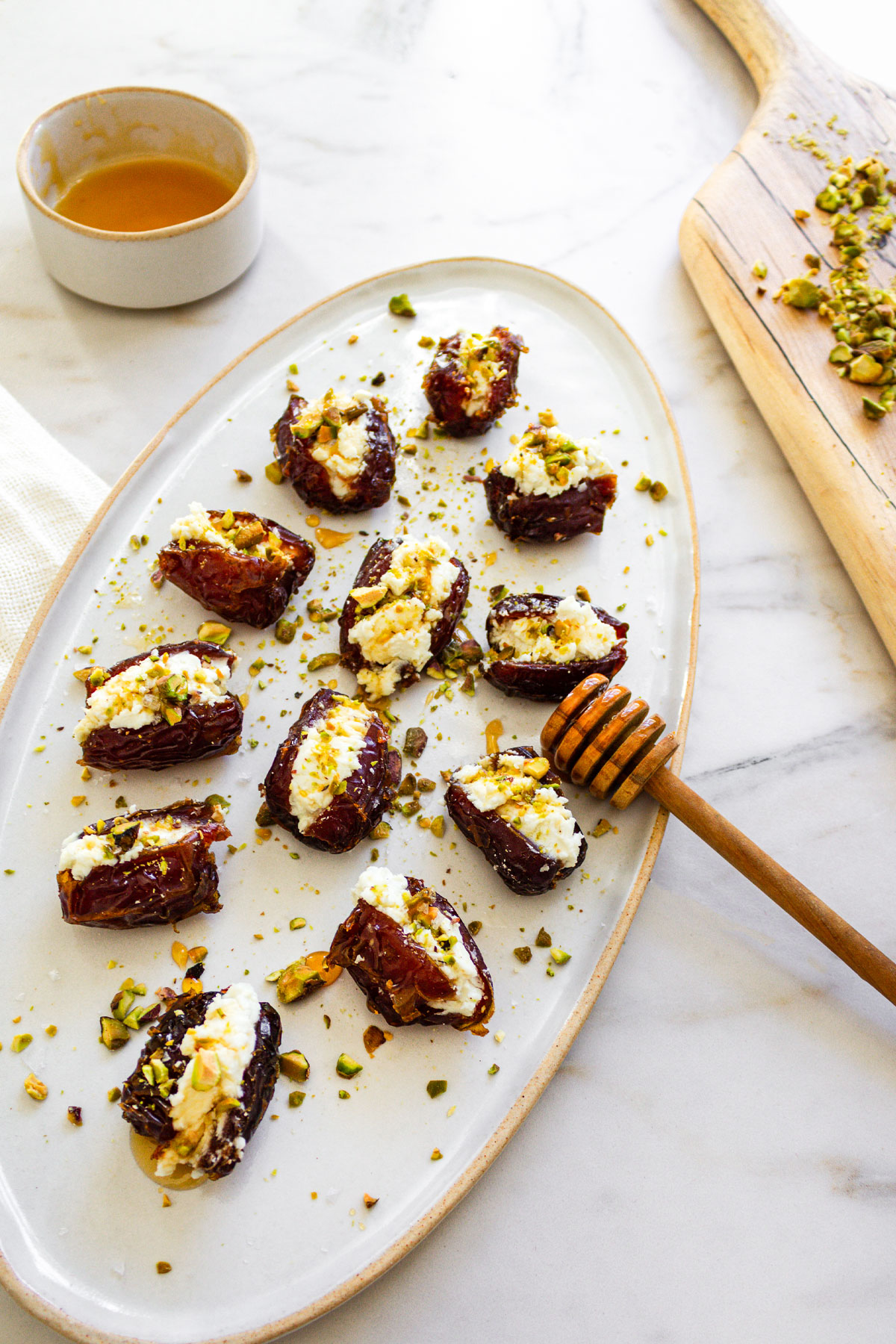 A white oval ceramic platter on a marble counter filled with Goat Cheese Stuffed Dates. The dates are drizzled with hot honey and topped with chopped pistachios which are scattered on the platter.