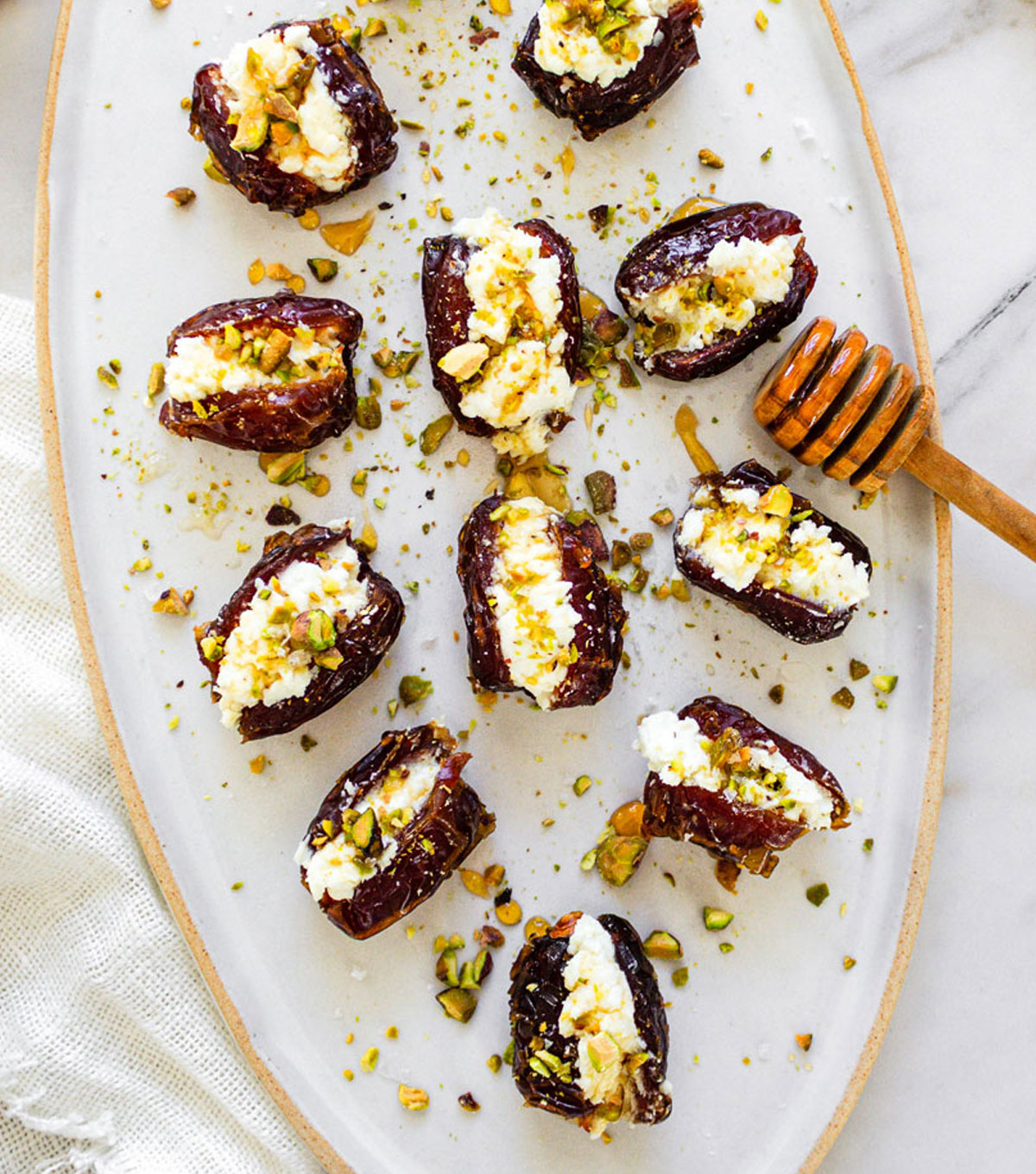 A white ceramic oval platter with Goat Cheese Stuffed Date appetizers drizzled with hot honey, with a honey dipper rested on the plate.
