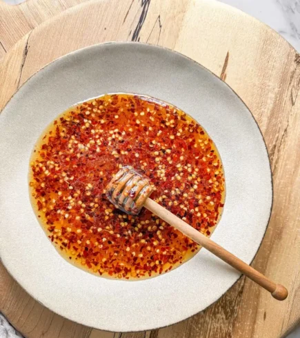 Hot honey infused with red chili peppers in a bowl on a wooden serving board, with a honey dipper in it.