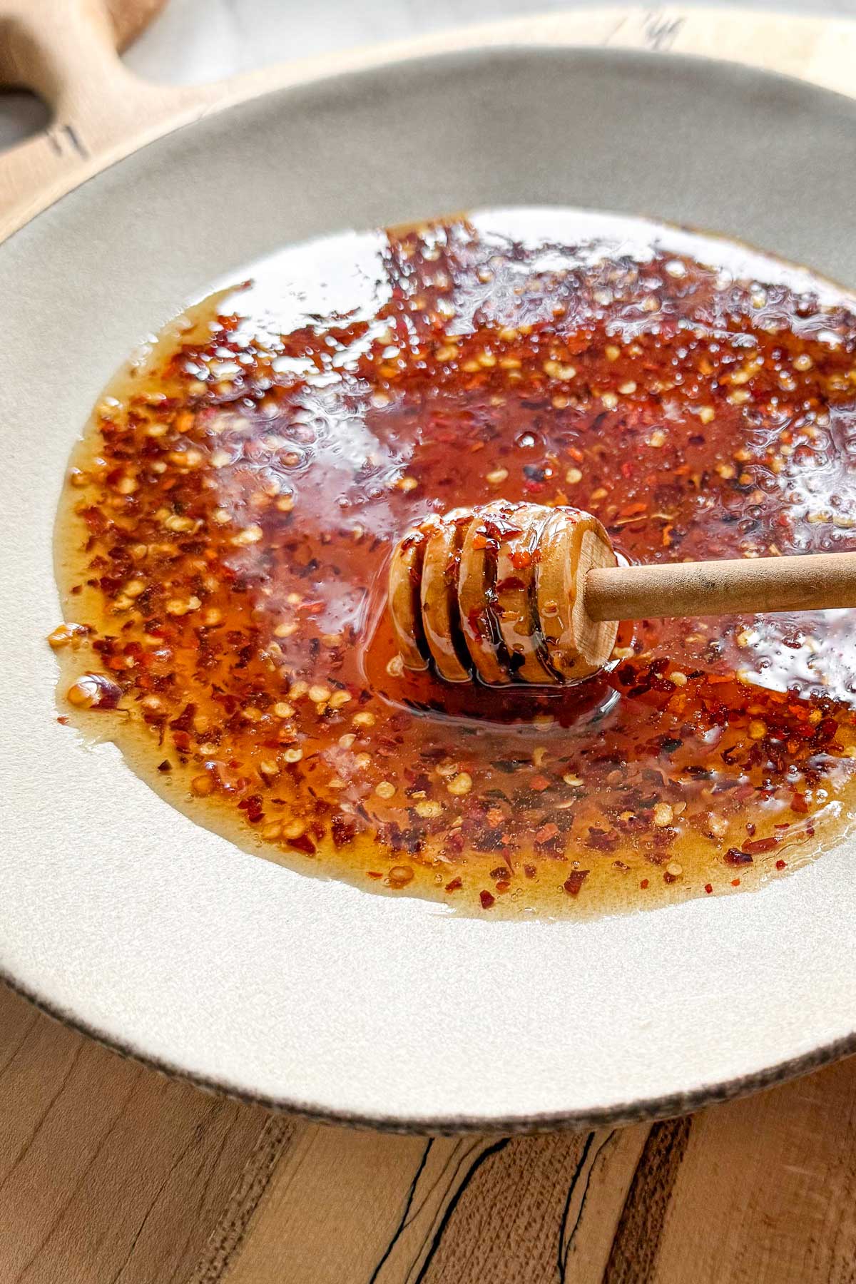 Hot honey infused with red chili peppers in a bowl on a wooden serving board, with a honey dipper in it.