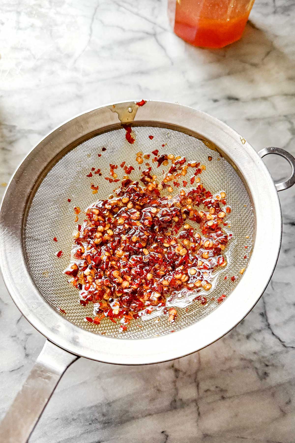 Hot honey infused with chili peppers being strained through a mesh sieve.