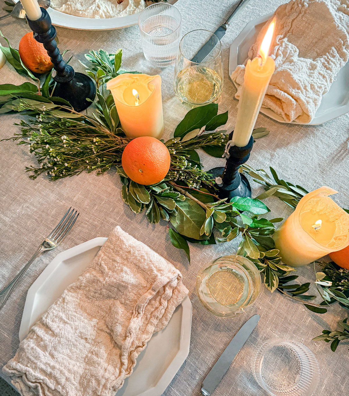 A dining table with an ivory linen tablecloth set with white plates and raw linen napkins. Candles and fresh greens and oranges line the center of the table.