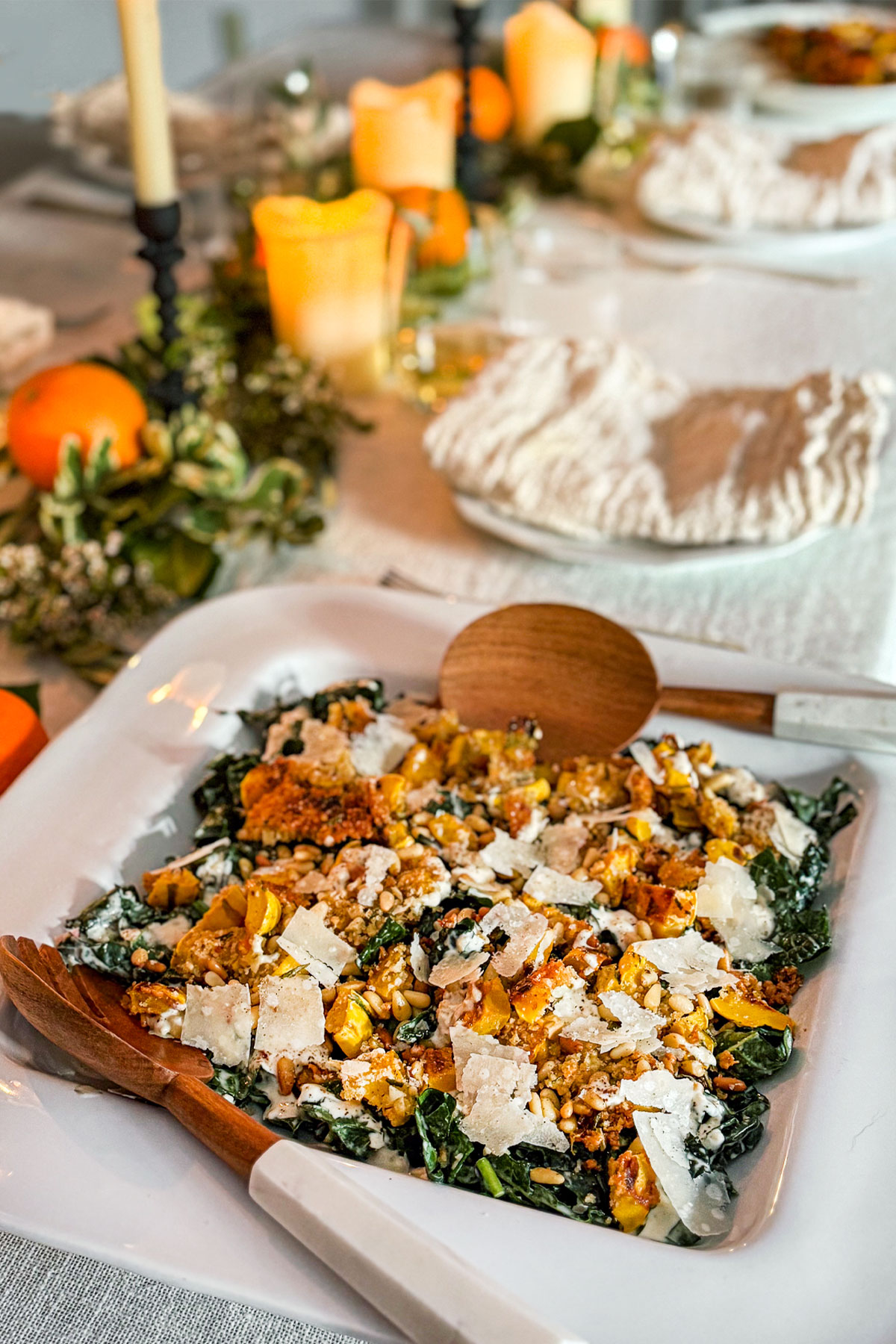 A dining table covered in an ivory linen tablecloth with a runner of candles, fresh greens, and citrus fruit. The table is set and a platter of Kale Caesar Salad topped with Roasted Delicata Squash with a crusted parmesan pine nut topping, dusted with shaved parmesan cheese sits in the foreground.