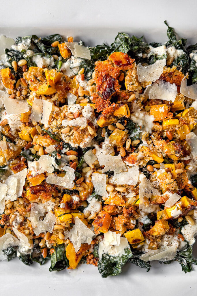 A close up of a platter layered with Kale Caesar Salad topped with Roasted Delicata Squash with a crusted parmesan pine nut topping, dusted with shaved parmesan cheese.