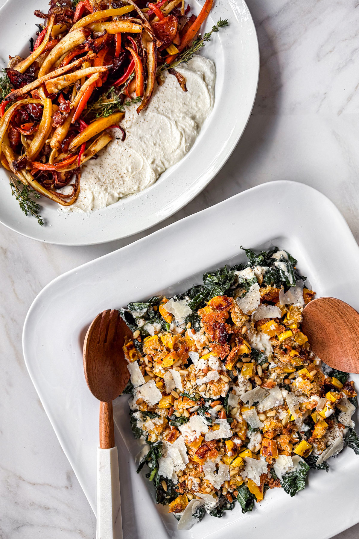 A marble counter with two serving platters side by side. One is a Kale Caesar Salad topped with Roasted Delicata Squash with a crusted parmesan pine nut topping, dusted with shaved parmesan cheese. The other is Roasted Parsnips & Carrots with Oranges & Whipped Feta.