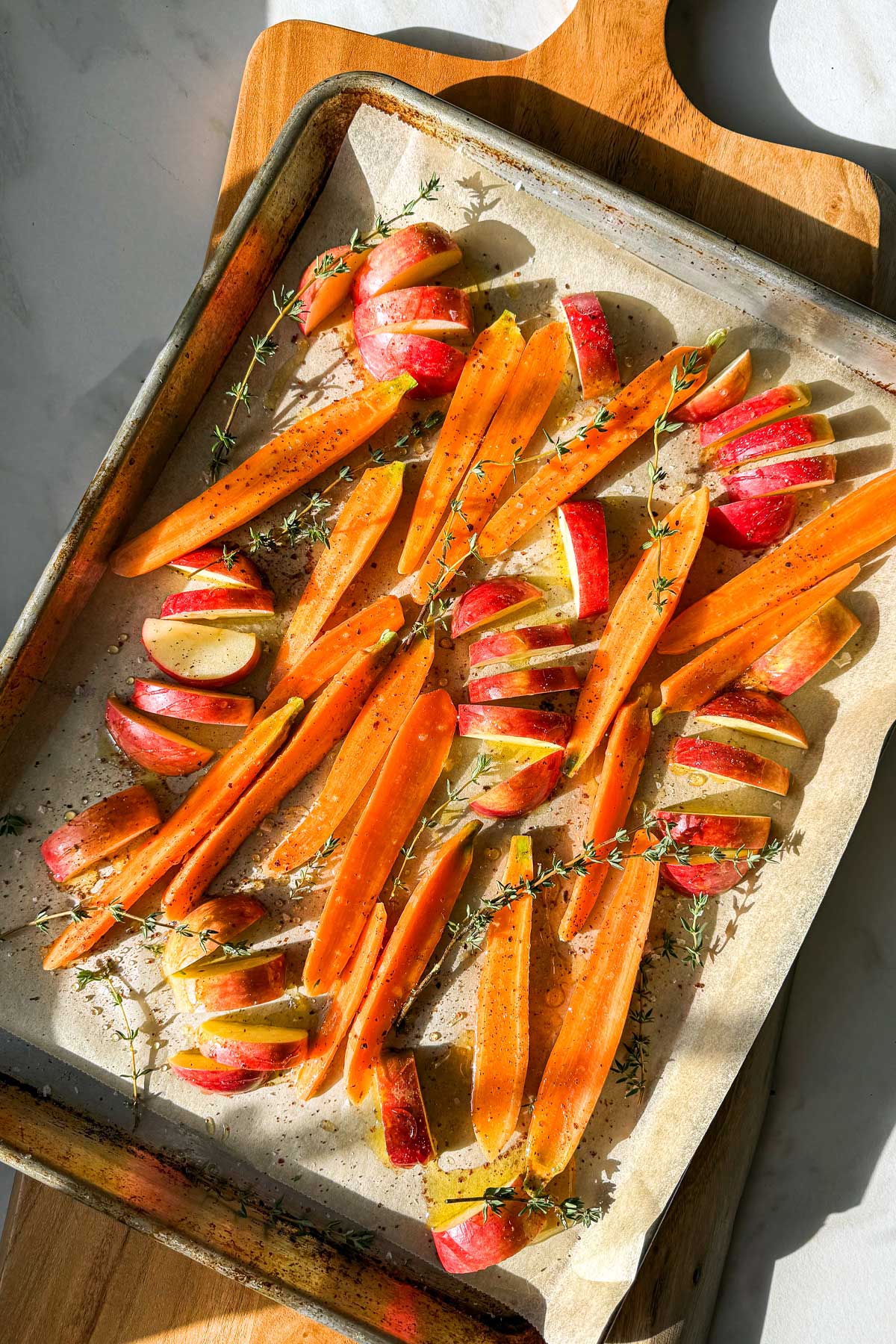 A baking sheet lined with parchment paper filled with sliced carrots and apples scattered with thyme sprigs and drizzled with olive oil, waiting to be roasted, shining in the bright winter light. 