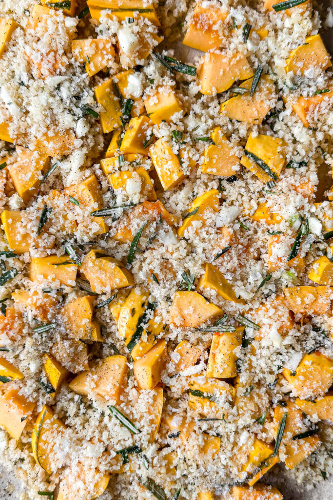 A close up of a baking tray, before baking, of cubed delicata squash covered with a breaded topping of panko breadcrumbs, parmesan cheese, and fresh rosemary.
