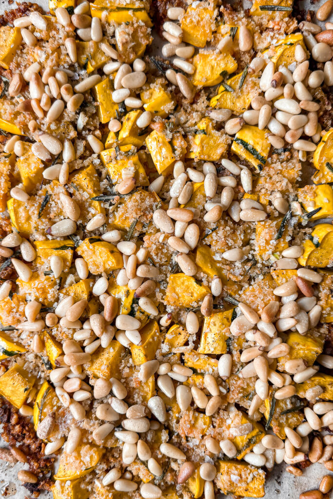 A close up of a baking tray out of the oven with roasted delicata squash crusted with panko, parmesan cheese, rosemary, and a layer of toasted pine nuts.