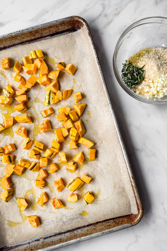 A sheet pan lined with parchment paper with a layer of cubed delicata squash drizzled with olive oil. Nect to it is a bowl of breading toppings.