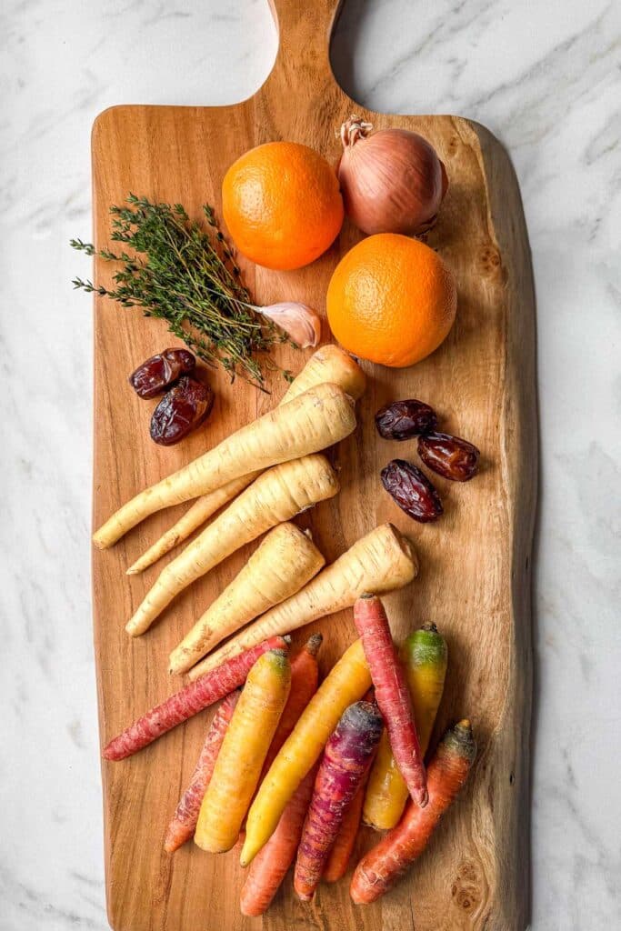 A large wooden serving board with the ingredients for Roasted Parsnips, & Carrots With Oranges & Whipped Feta laid out: Oranges, Parsnips, Rainbow Carrots, Dates, Garlic, Onion, and Thyme