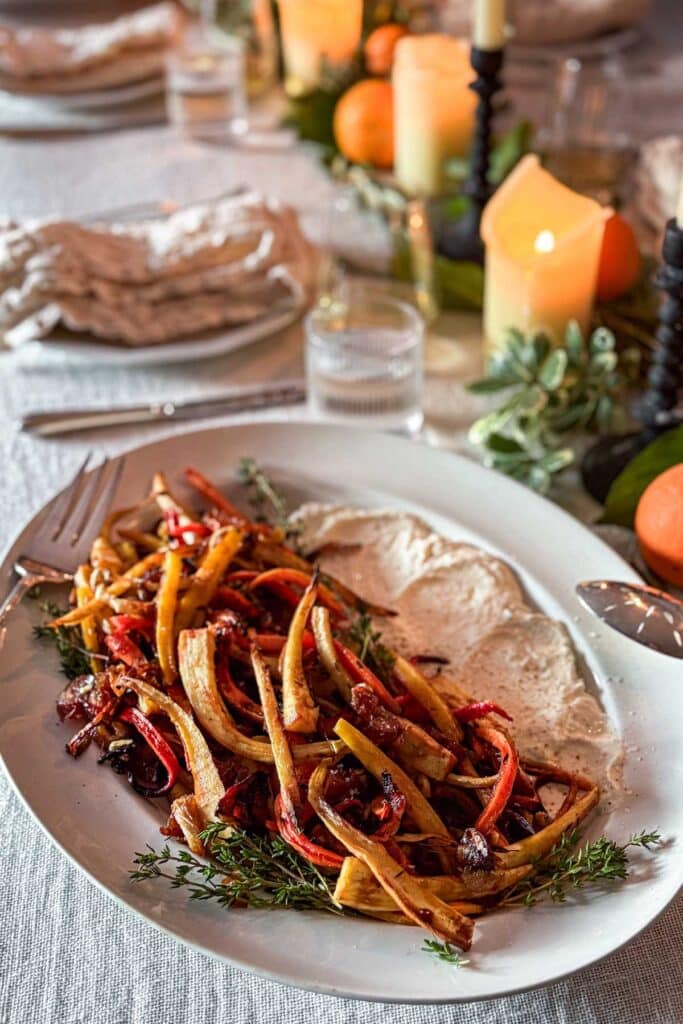 A set dinner table with an ivory linen tablecloth and a centerpiece of greens, candles, and citrus and in the foreground a serving platter with a generous smear of whipped feta finished with caramelized parsnips, carrots, and oranges with sprigs of fresh thyme tucked in.