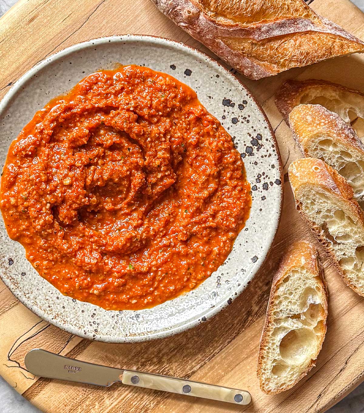 A maple serving board with a low speckled dish filled with chunky warm Romesco sauce, surrounded by fresh baguette for dipping.