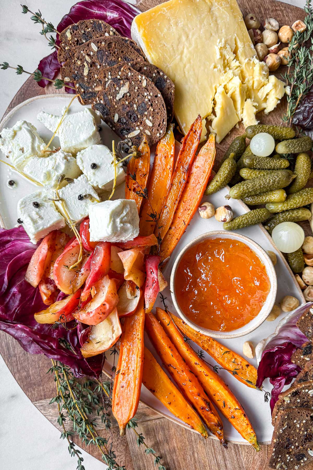A close-up image of a winter cheeseboard. On a wooden board, there is a slab of Welsh-style cheddar cheese, pickled cornichons and onions, roasted apples and carrots, a marinated sheep's and goat's milk cheese. Surrounding a small natural ramekin of marmalade. 