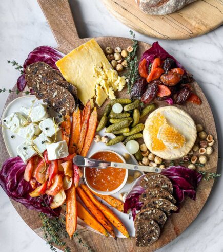 A large round wooden cheese board with winter cheeses and accompanimentsL Marinated Sheep's & Goat's milk cheese, Welsh cheddar, and Willoughby washed rind cheese with roasted apples and carrots, cornichons, dates, and hazelnutes with seeded crackers.