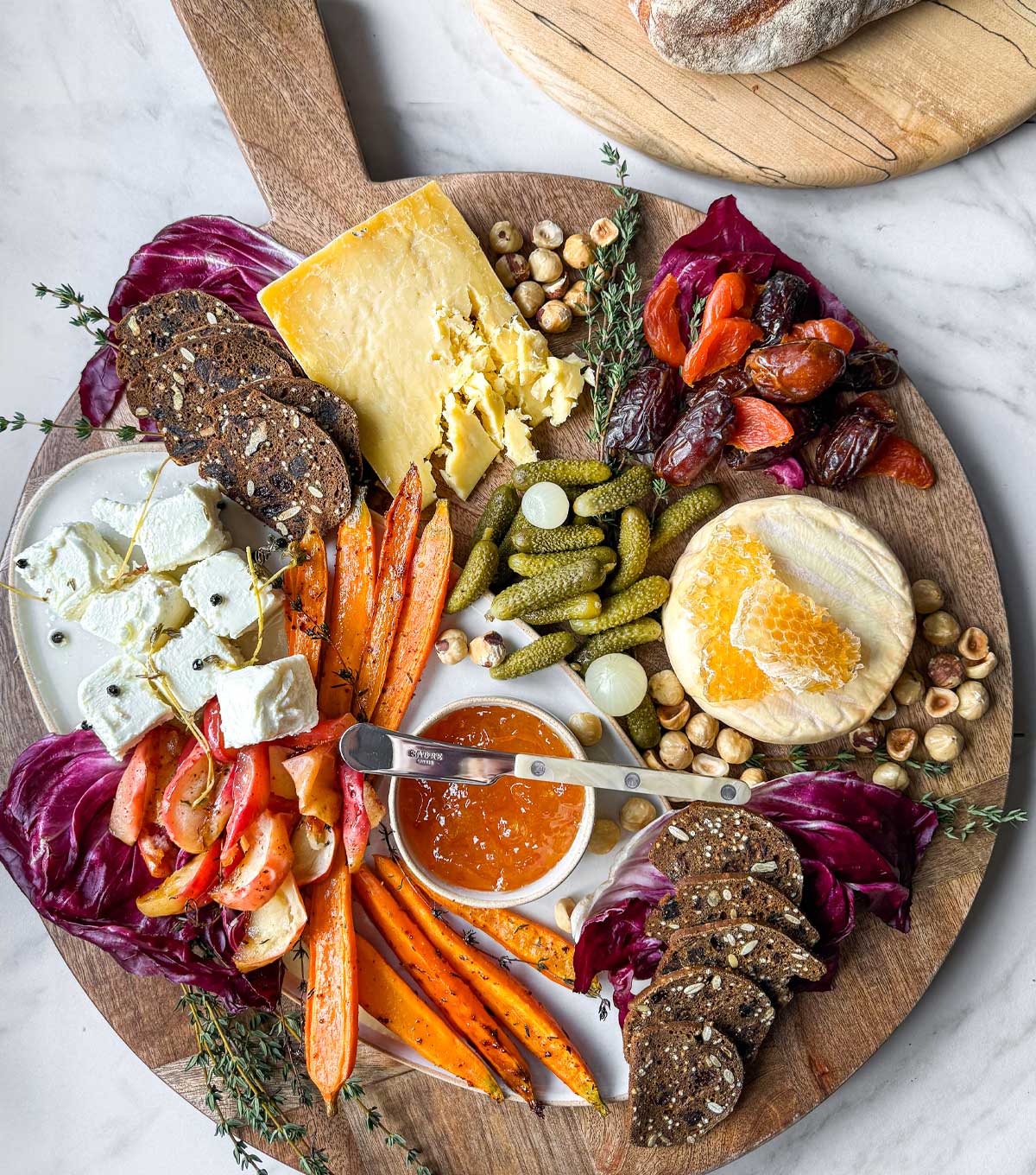 A large round wooden cheese board with winter cheeses and accompanimentsL Marinated Sheep's & Goat's milk cheese, Welsh cheddar, and Willoughby washed rind cheese with roasted apples and carrots, cornichons, dates, and hazelnutes with seeded crackers.