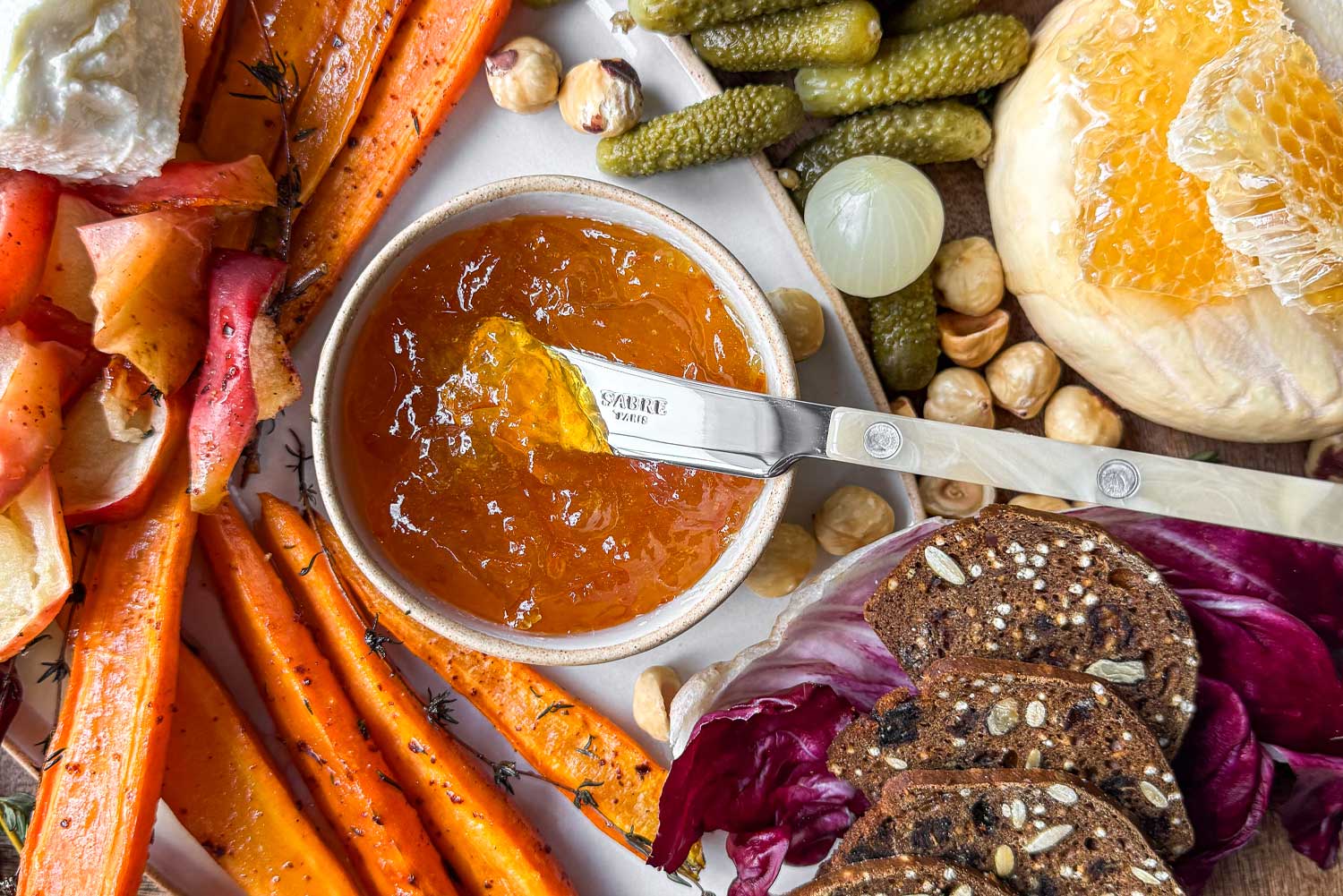 A close-up of a ramekin of marmalade with a Sabre Paris cheese spreader in it. Alongside washed rind cheeses with honeycomb, seeded crackers, cornichons, hazelnuts, and roasted apples and carrots. 
