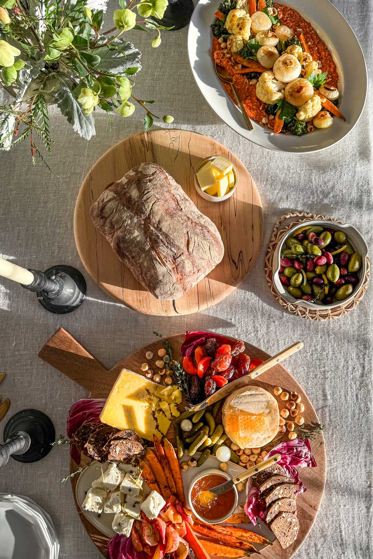 A styled table at a winter wine and cheese party. The table is covered with an ivory linen cloth and filled with grazing and sharing platters. There is a winter cheese board on a large round wooden board. A board with ciabatta and butter. A serving bowl with warm olives and citrus. And a serving platter with winter roasted vegetables and Romesco sauce. Interspersed with these are candles and a winter arrangement of hellebores. 