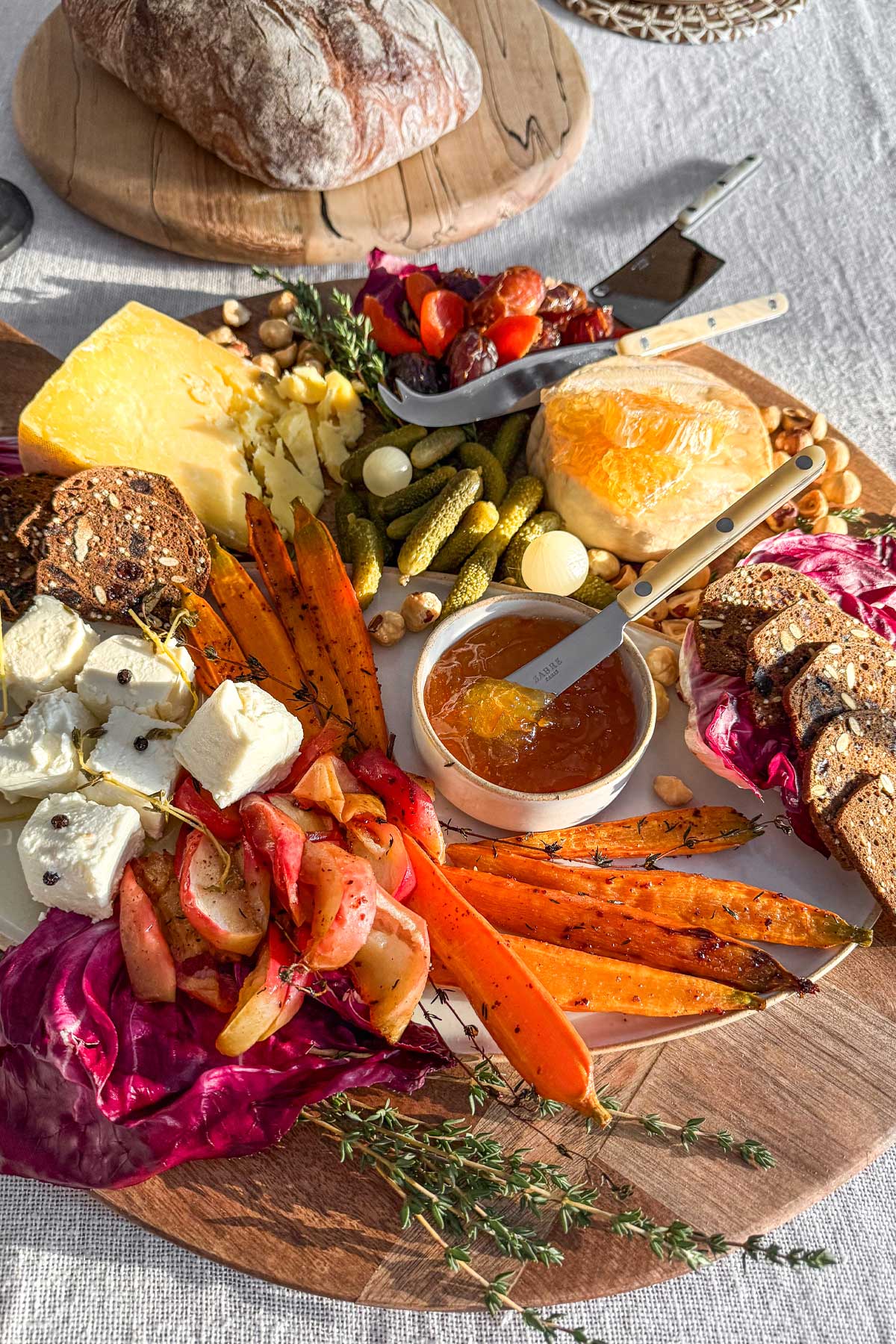 A close-up image of a winter cheese board at a winter wine and cheese party. On an ivory linen tablecloth, there is a large round board filled with seasonal winter cheeses, pickled elements, roasted apples and carrots, and marmalade. 