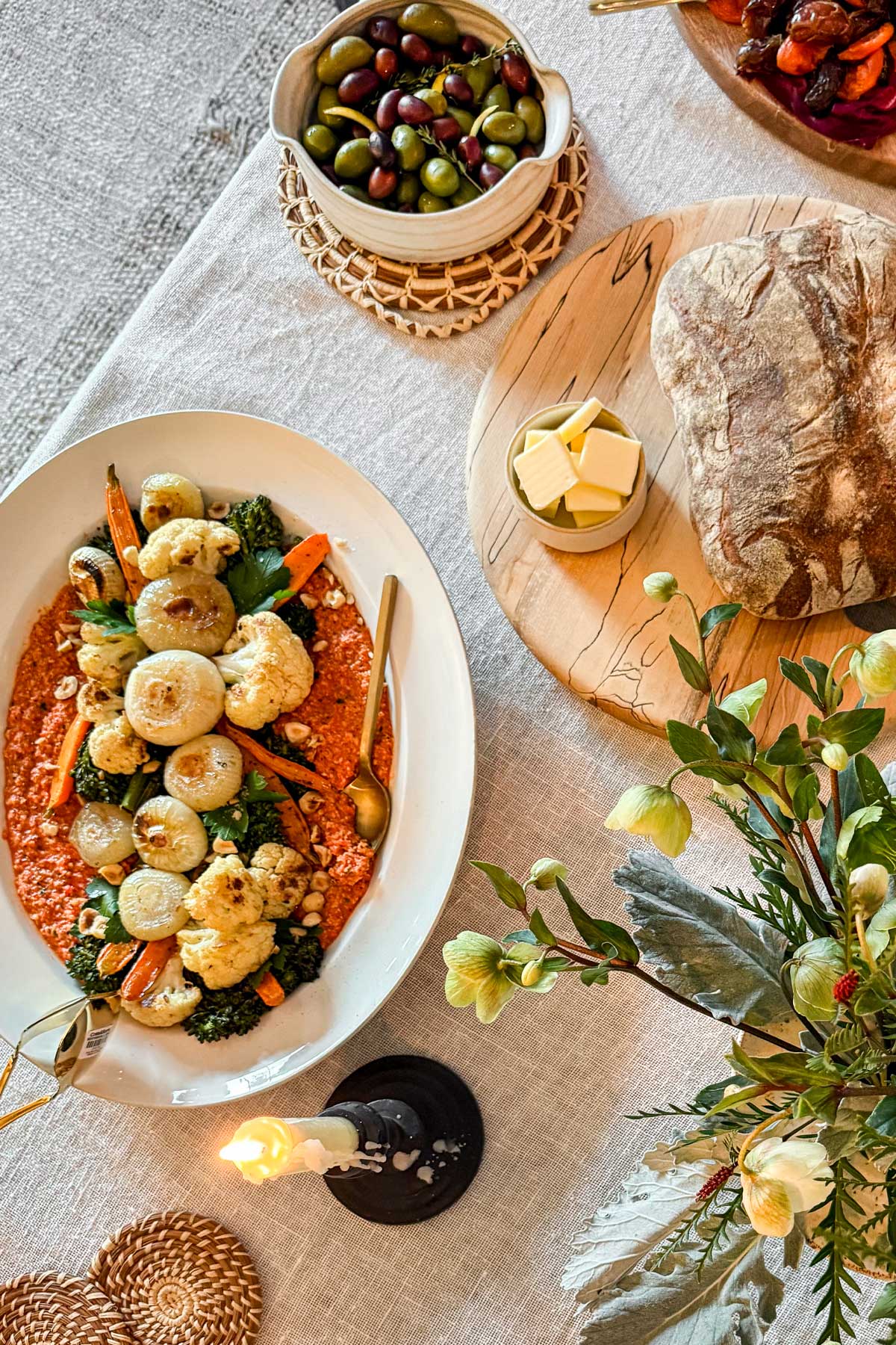 A dining table with an ivory linen tablecloth is set with a platter of romesco sauce and roasted winter vegetables alongside thick rustic bread and marinated olives.