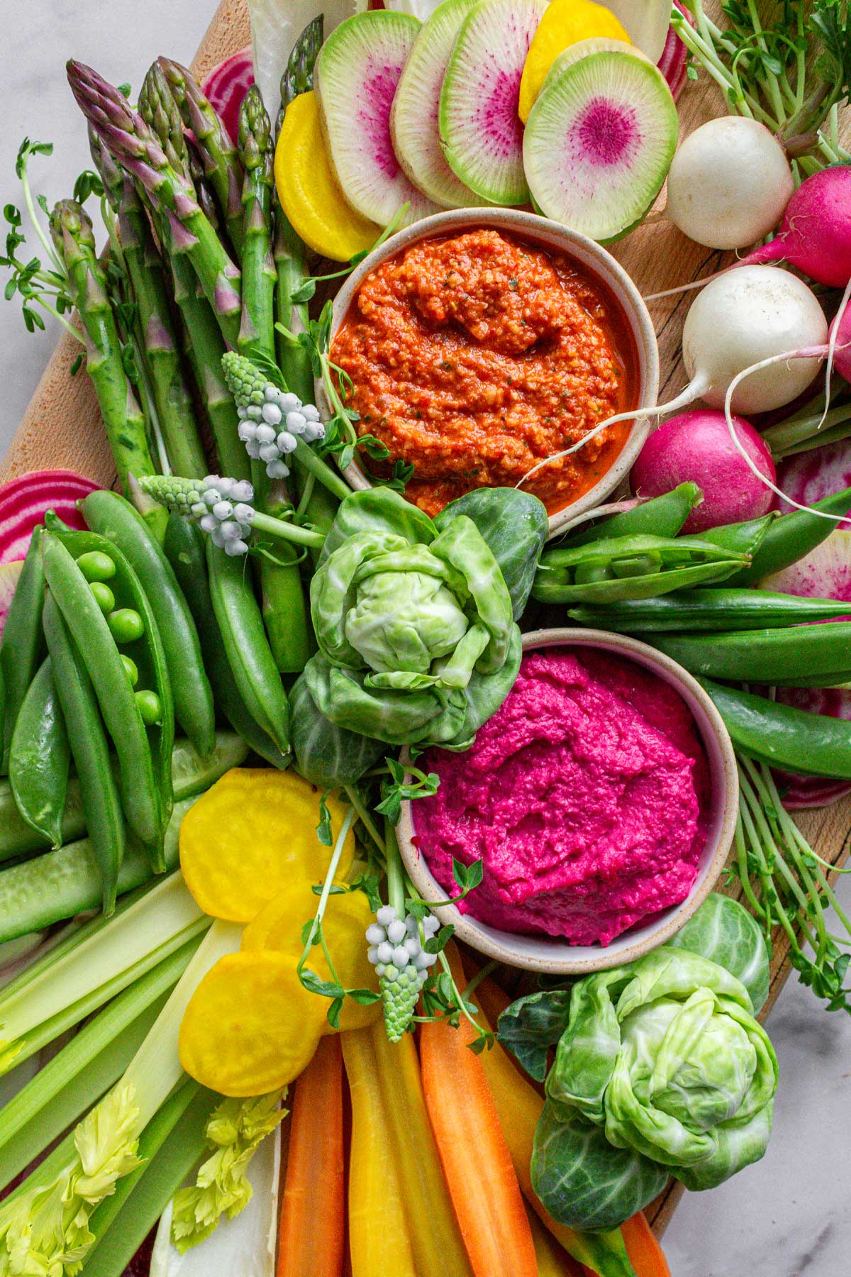 A spring crudité board layered with asparagus, peas, radishes, carrots, around a ramekins of romesco sauce and beet hummus.