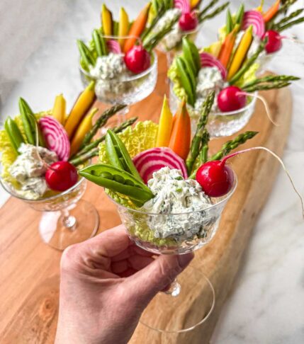 Garden crudité goblets in small stemmed glasses, filled with garlic herb dip and arranged with asparagus, snap peas, radishes, watermelon radish slices, and carrots, styled on a wooden board for spring entertaining