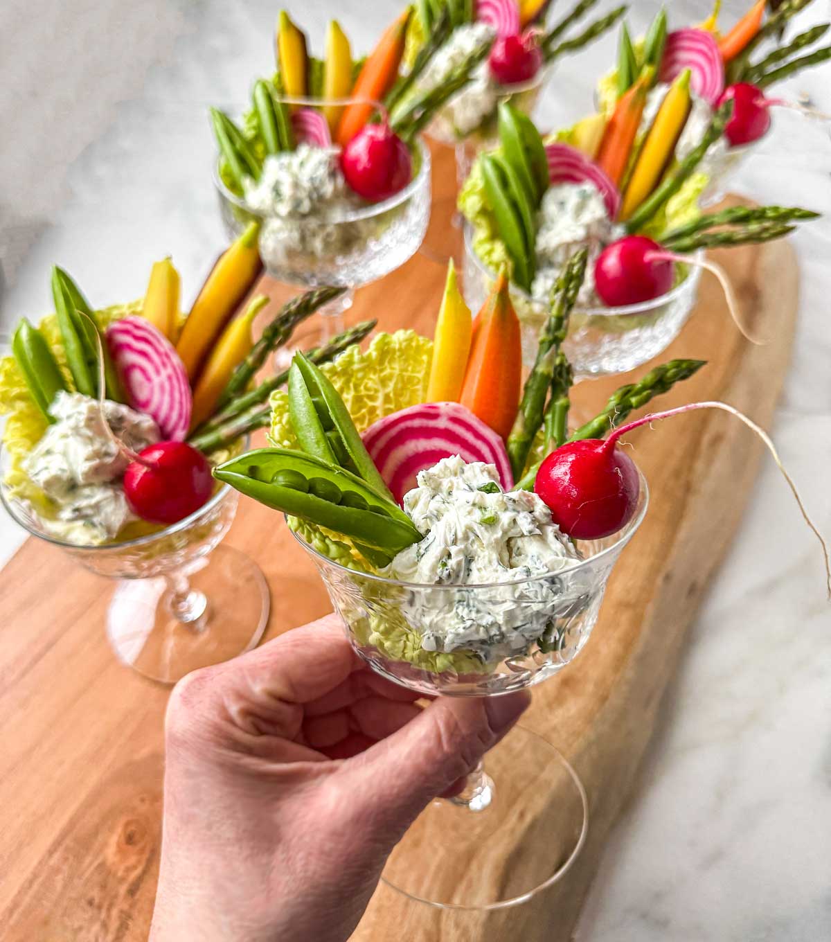 Garden crudité goblets in small stemmed glasses, filled with garlic herb dip and arranged with asparagus, snap peas, radishes, watermelon radish slices, and carrots, styled on a wooden board for spring entertaining