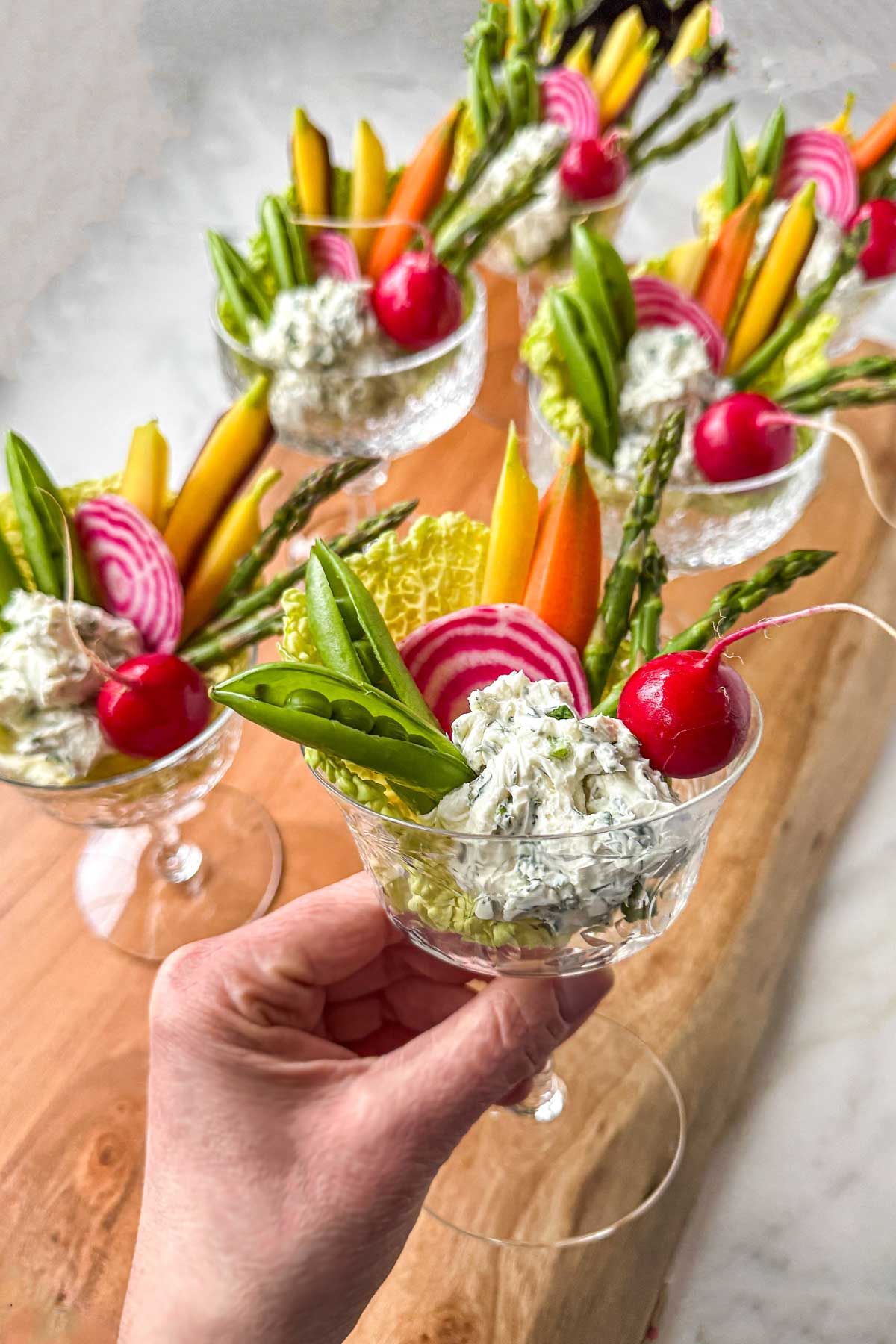 Garden crudité goblets in small stemmed glasses, filled with garlic herb dip and arranged with asparagus, snap peas, radishes, watermelon radish slices, and carrots, styled on a wooden board for spring entertaining