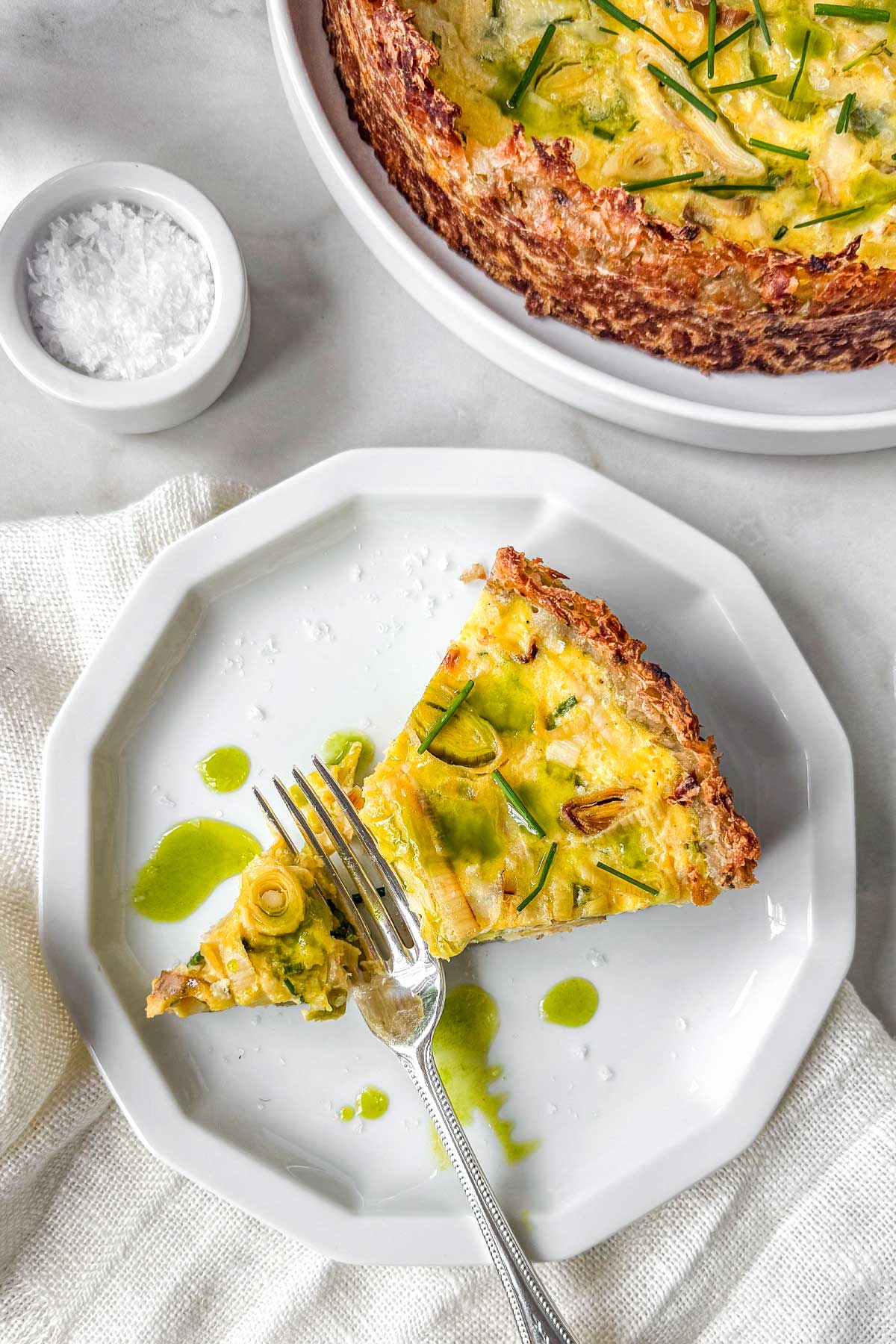 Hashbrown crust quiche with leeks and Gruyère topped with chives and drizzled chive oil, served on a marble table with a sliced piece on a plate