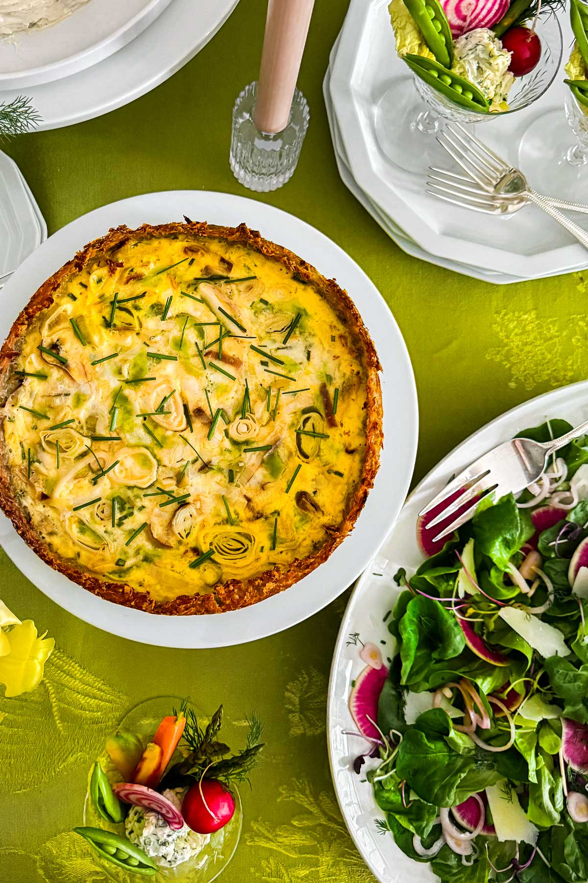 Hashbrown crust quiche with leeks and chives served on a spring brunch table with green salad, crudité, and seasonal vegetables