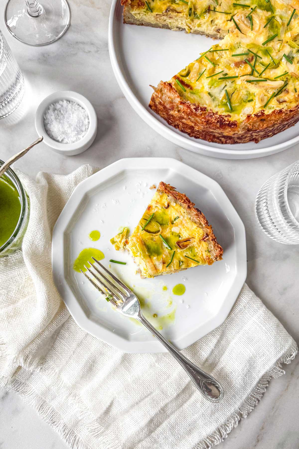 Hashbrown crust quiche with leeks and Gruyère topped with chives and drizzled chive oil, served on a marble table with a sliced piece on a plate