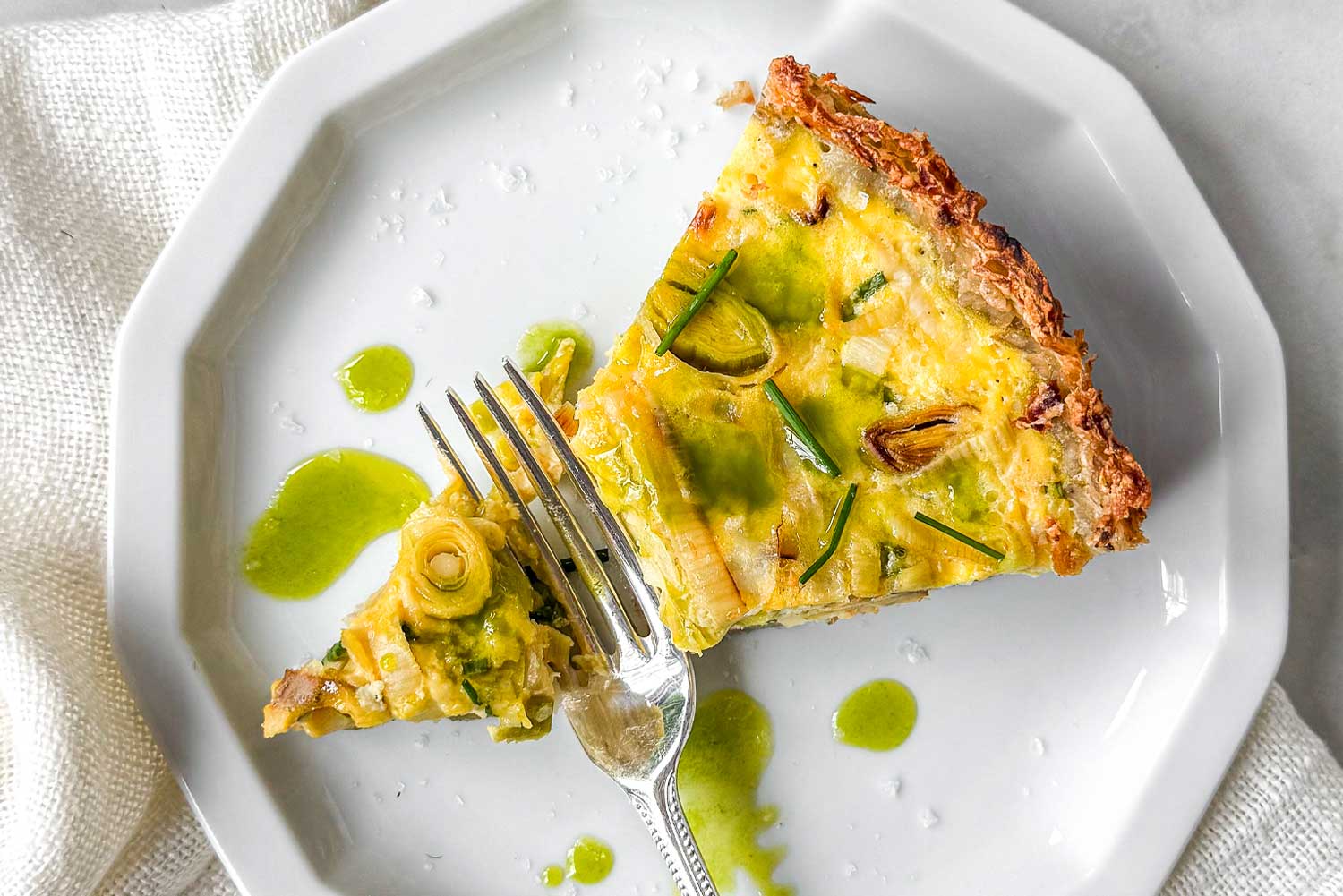 Hashbrown crust quiche with leeks and Gruyère topped with chives and drizzled chive oil, served on a marble table with a sliced piece on a plate